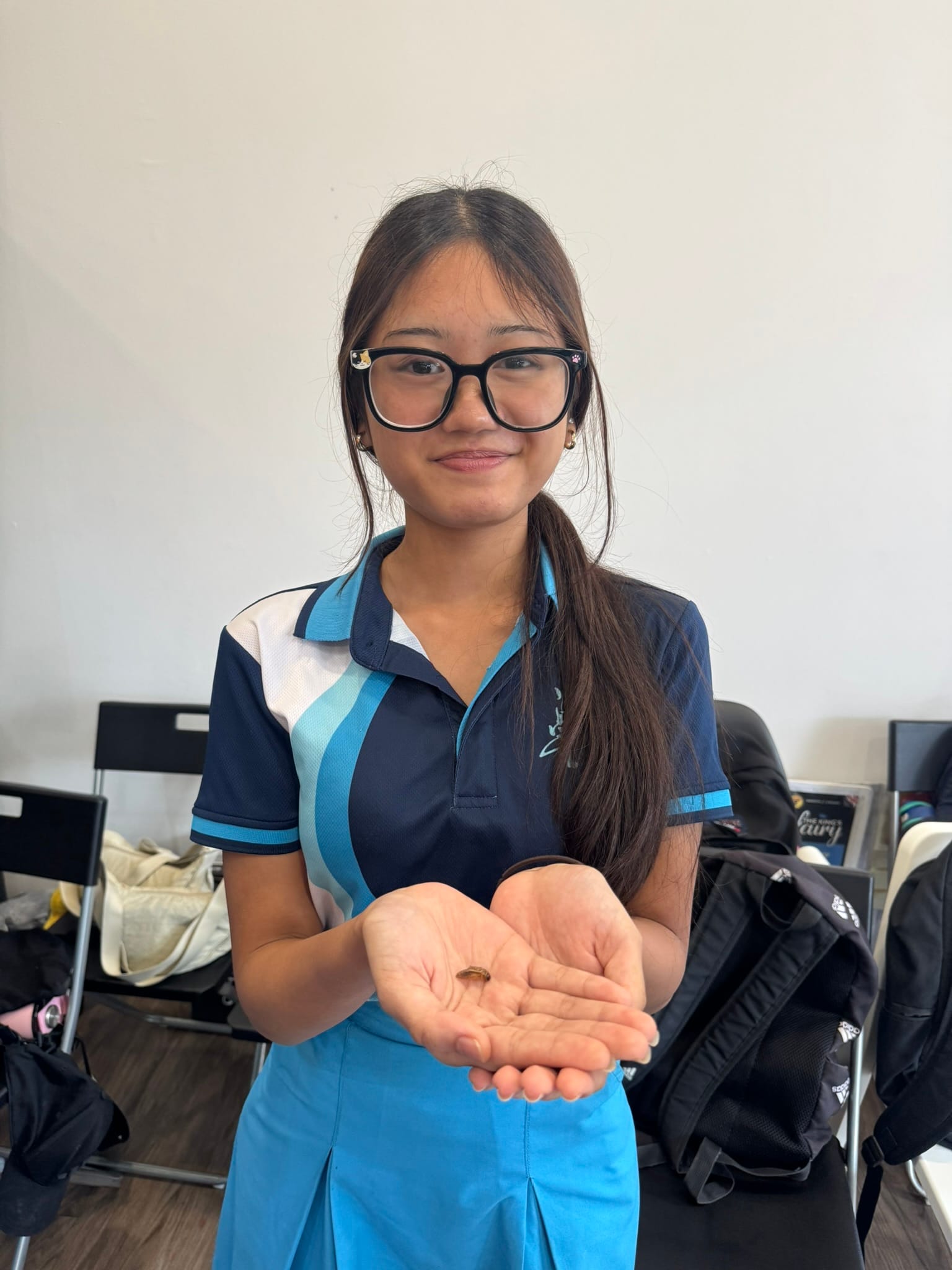 A smiling female student in school uniform holding out her cupped hands to display a small food sample or ingredient during a practical lesson.